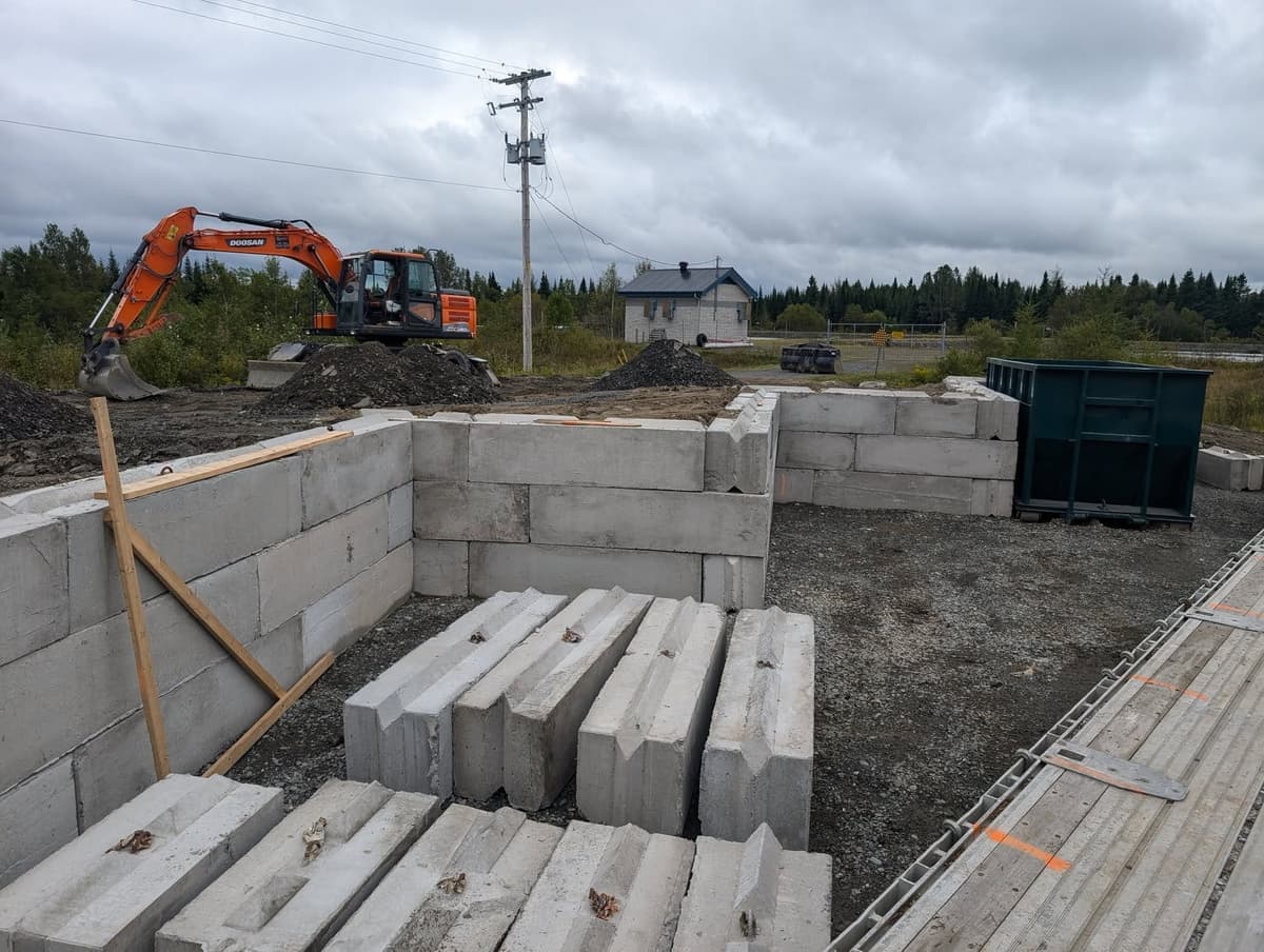 Blocs de béton préfabriqués posés sur chantier avec excavatrice orange en arrière-plan — A.L.O Pomerleau