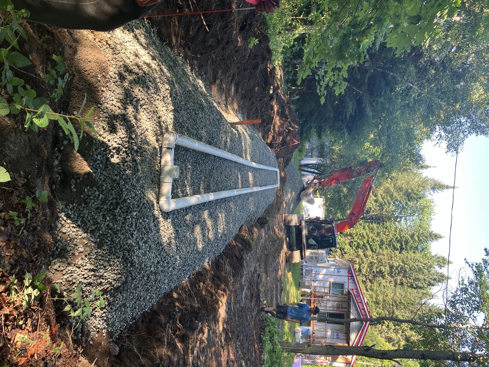 Pelle hydraulique rouge et tranchée de drainage avec conduites PVC sur lit de gravier devant une résidence à Tring-Jonction