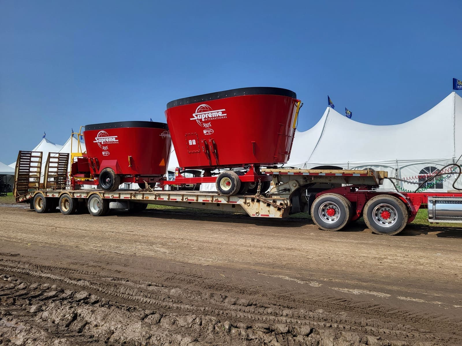 Deux gros mélangeurs agricoles rouges chargés sur fardier dans un champ, près de tentes blanches