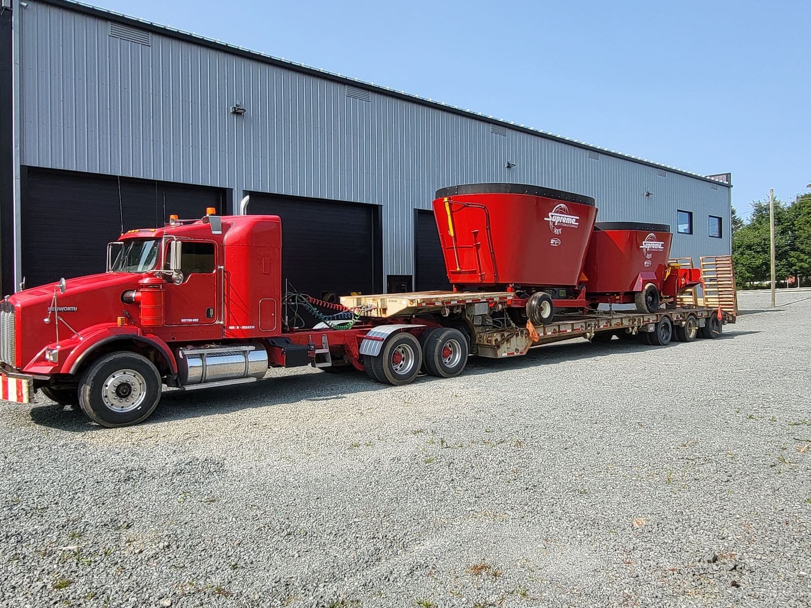 Camion fardier transportant deux mélangeurs agricoles rouges devant un entrepôt
