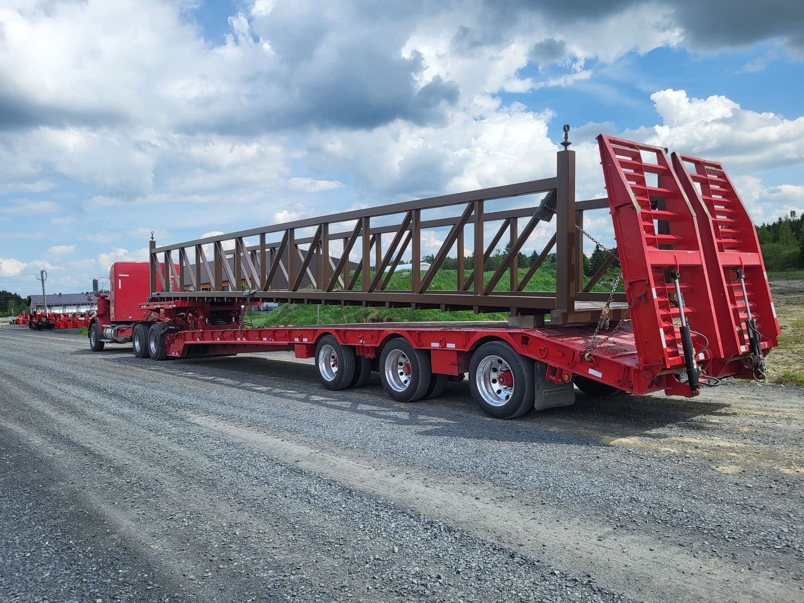 Camion fardier transportant une longue charpente métallique brune en treillis sur un terrain de gravier