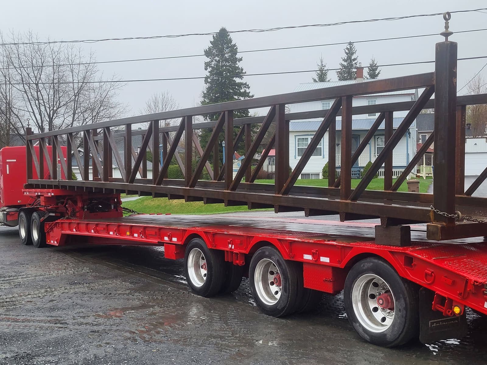 Vue arrière d'un fardier transportant une charpente métallique brune devant des résidences