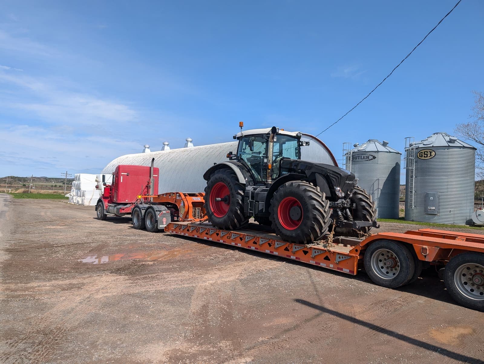 Camion fardier transportant un gros tracteur agricole devant des silos à grain